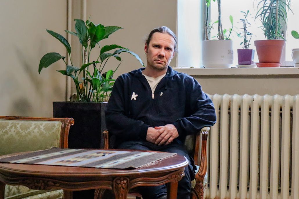 Kuvassa miesoletettu istuu puisen pöydän ääressä. Pöydällä on pöytäliina, ja taustalla näkyy viherkasveja.