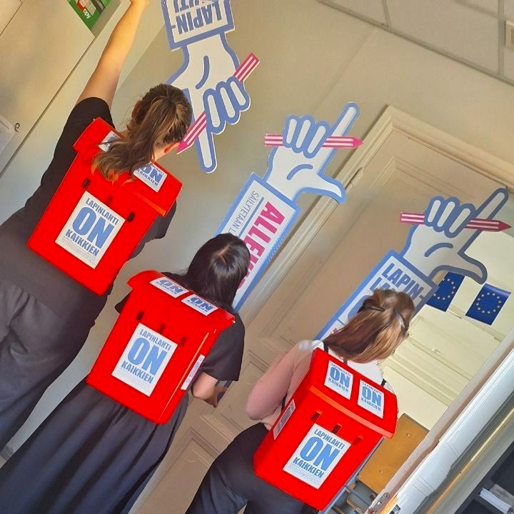 Kolme naista seisoo selkä kameraan päin. Heillä on selässään punaiset Lapinlahti-tarroilla koristellut postilaatikot