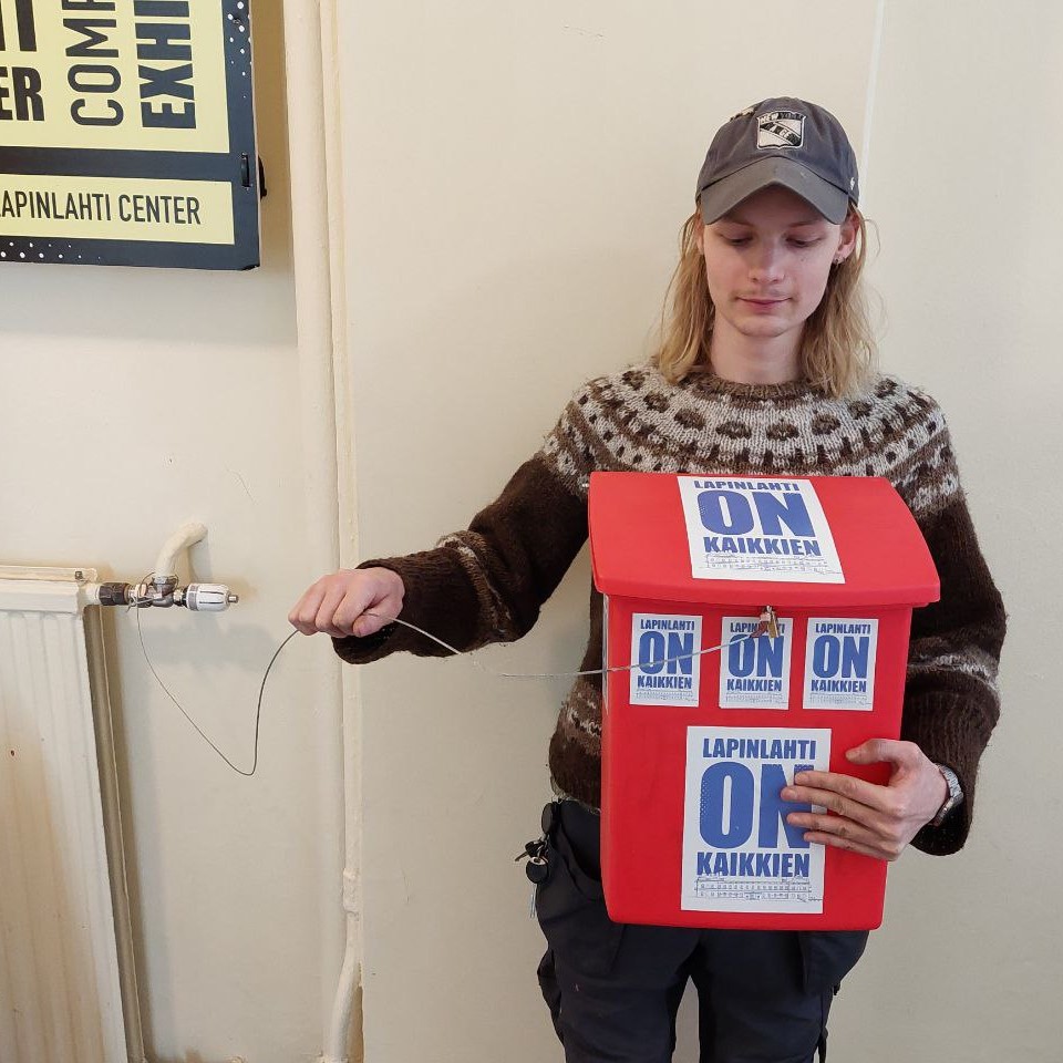 Nuori mies pitelee kiinni vaijerista ja hänellä on punainen tarroin koristeltu postilaatikko sylissään.