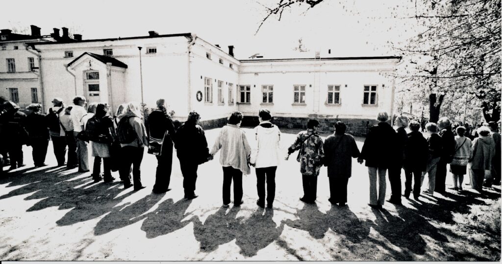 Mustavalkoisessa kuvassa ihmiset pitävät toisiaan kädestä päärakennuksen ympärillä.
