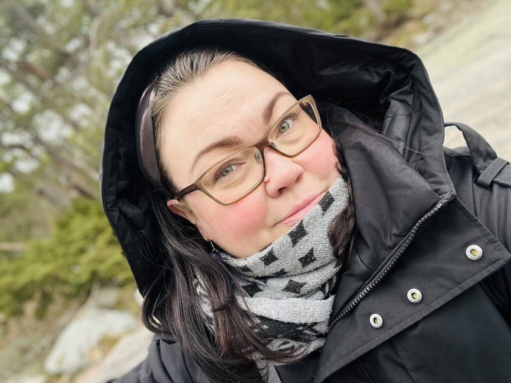 Kuvassa on nainen etualalla, huppu päässään. Taustalla metsää.
