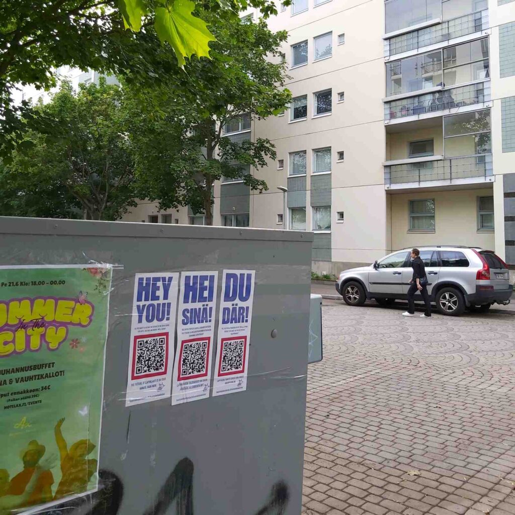 Kuva sähkökaapista, johon on teipattu kolmella kielellä julisteet, joissa on QR-koodi ja huudahdus "Hei sinä!"