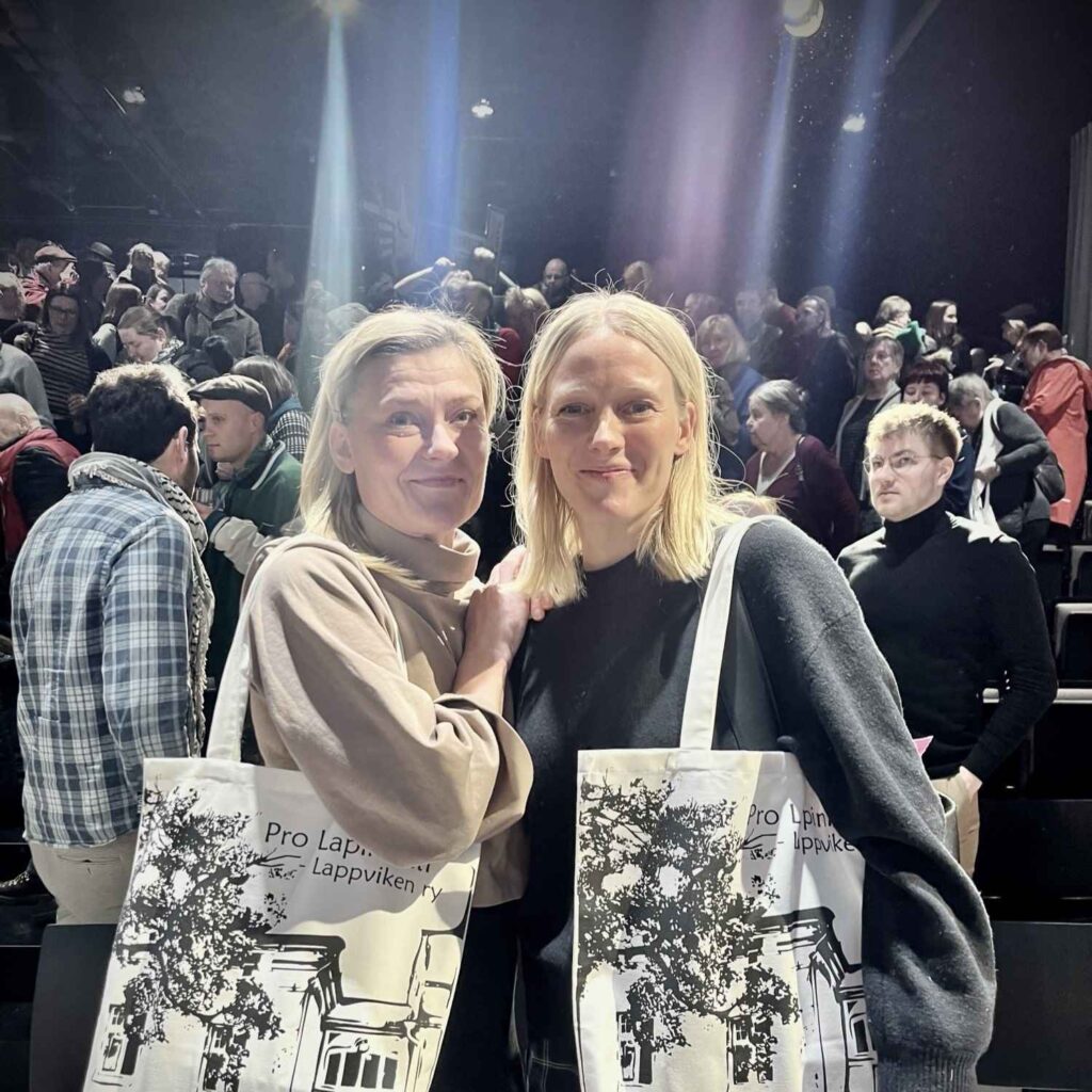 Kaksi naista hymyilee kameralle. Heillä on olillaan suuret vaaleat Pro Lapinlahti -kangaskassit. Tummalla taustalla on paljon ihmisiä ja värivaloja.