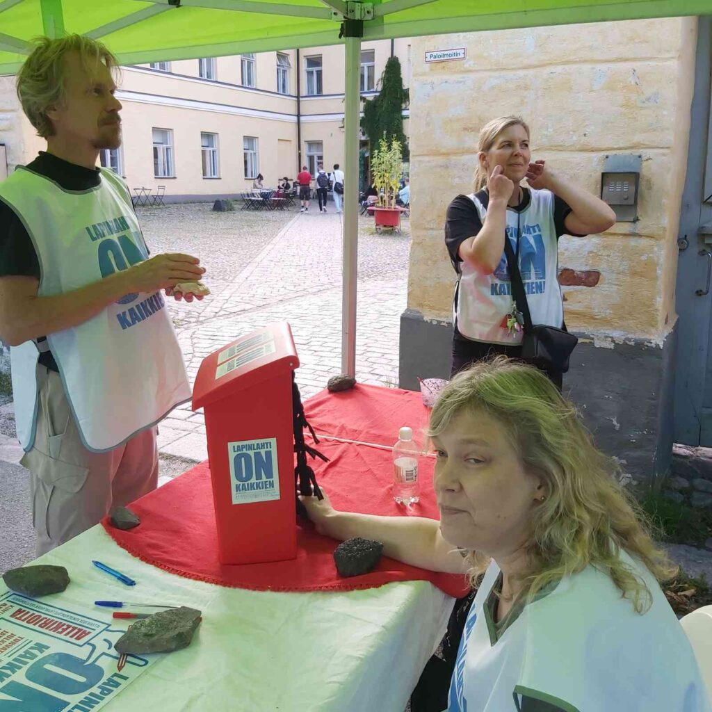 Kolme henkilöä ovat vihreän teltan alla ja katsovat vähän eri suuntiin. Pöydällä on punainen postilaatikko