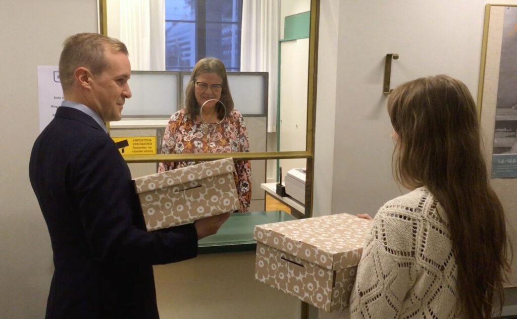 Kaksi henkilöä ojentavat pahvilaatikot tiskin yli kirjaamon työntekijälle