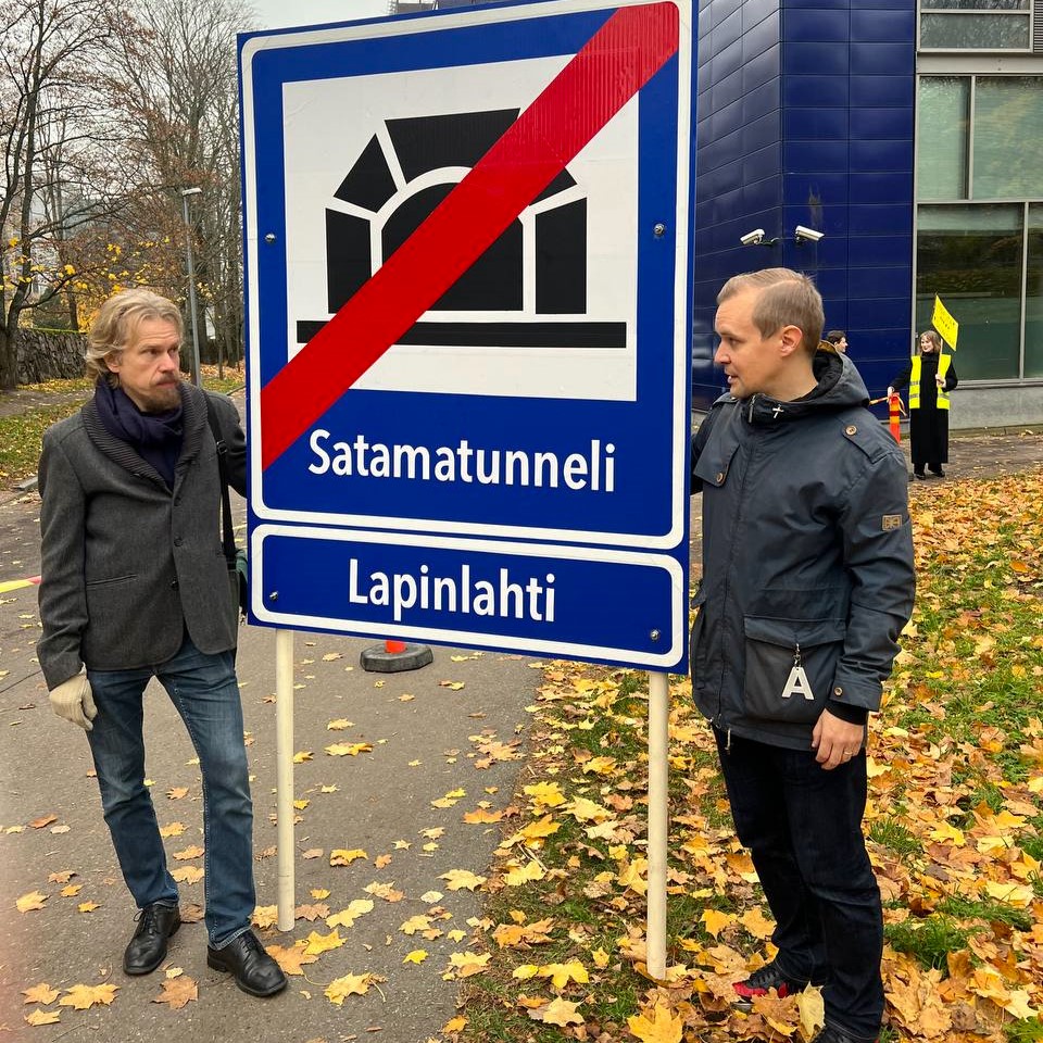 Kaksi miestä pitelevät suurta liikennemerkkiä, jossa satamatunnelin yli on vedetty paksu punainen viiva